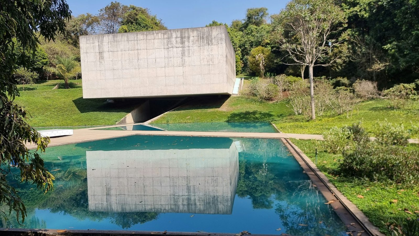 A raw concrete box cantilevers above a rectangular turquoise pool set in lawns and trees, its underside mirrored in the water