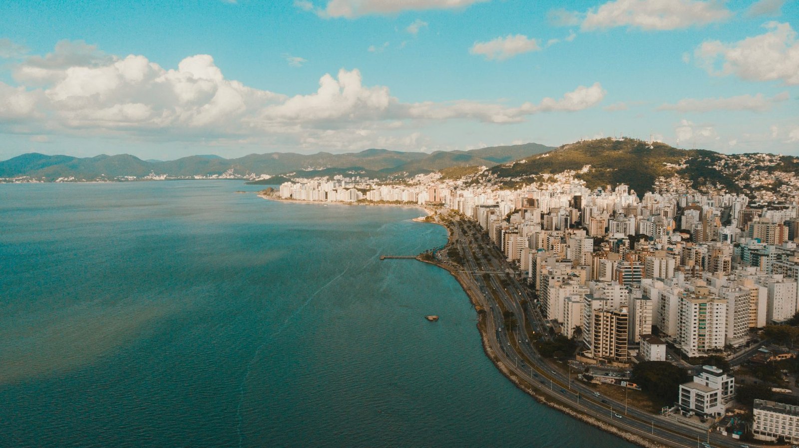 Aerial view of a dense row of white apartment towers following a curved shoreline road beside calm green water, with hills behind