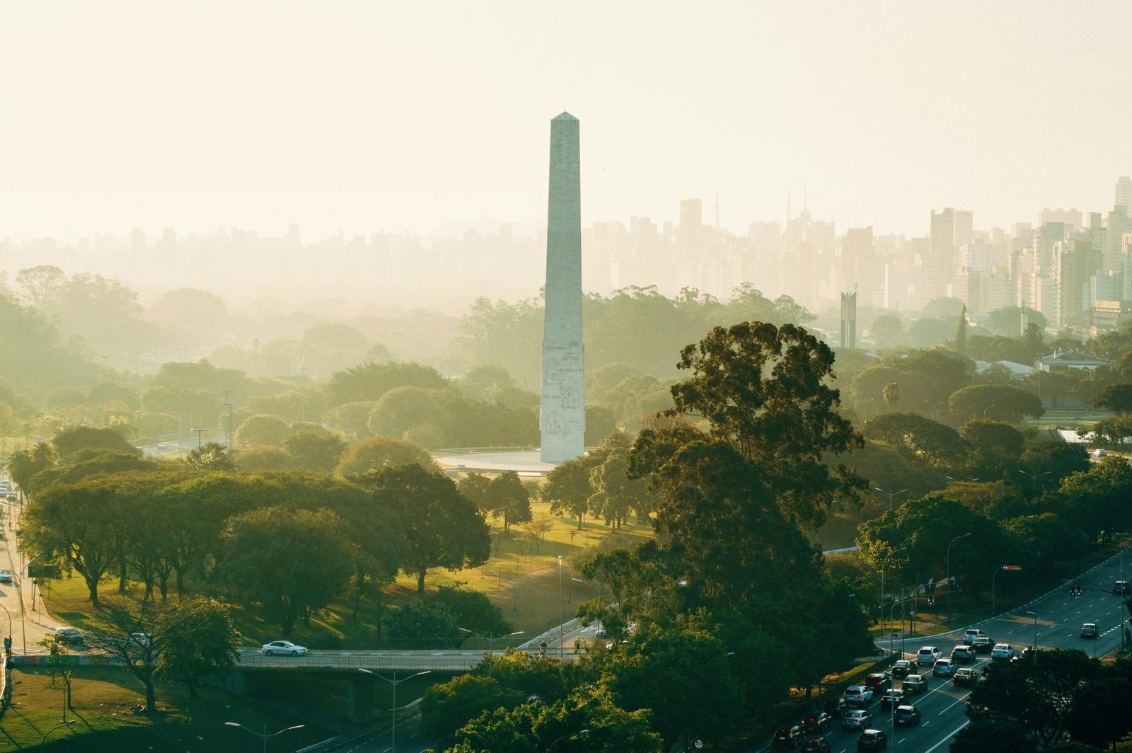 A tall stone obelisk rises from misty parkland and trees in golden light, with a wall of towers faint in the distance