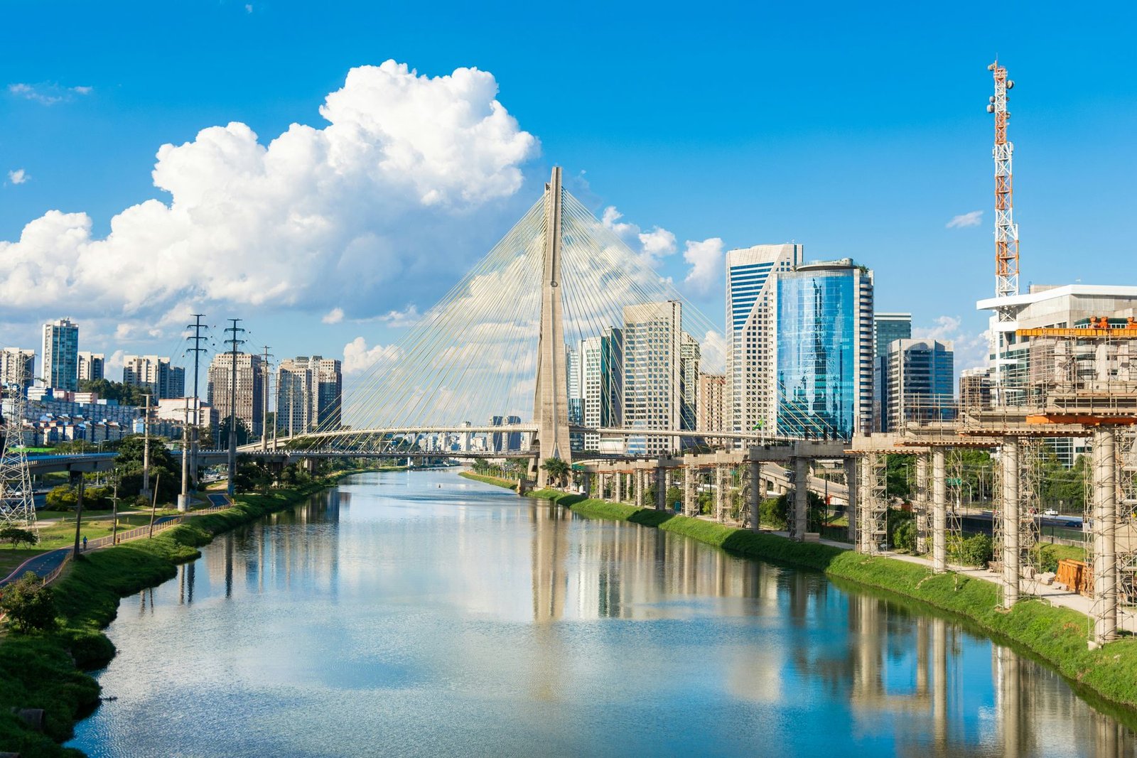 A cable-stayed bridge with an X-shaped mast crosses a calm river that reflects glass office towers and white clouds