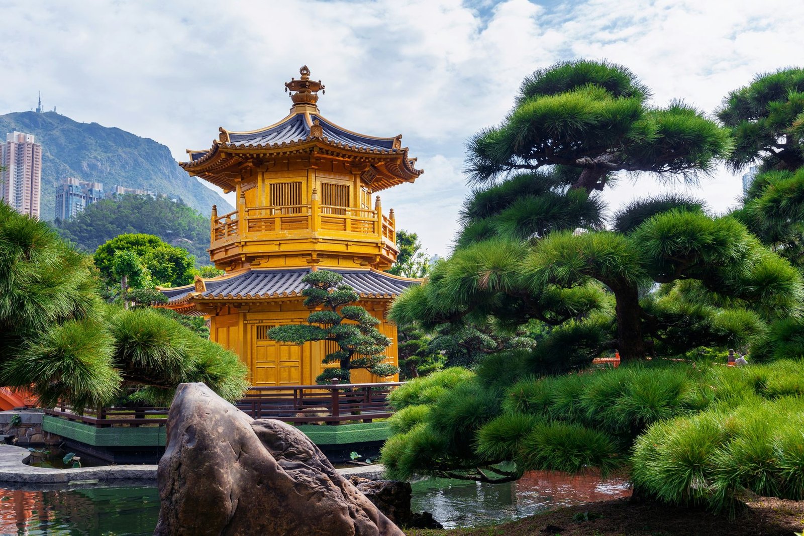 A two-tiered golden pavilion stands among clipped pines and rocks beside a pond, with a hillside and tower blocks behind