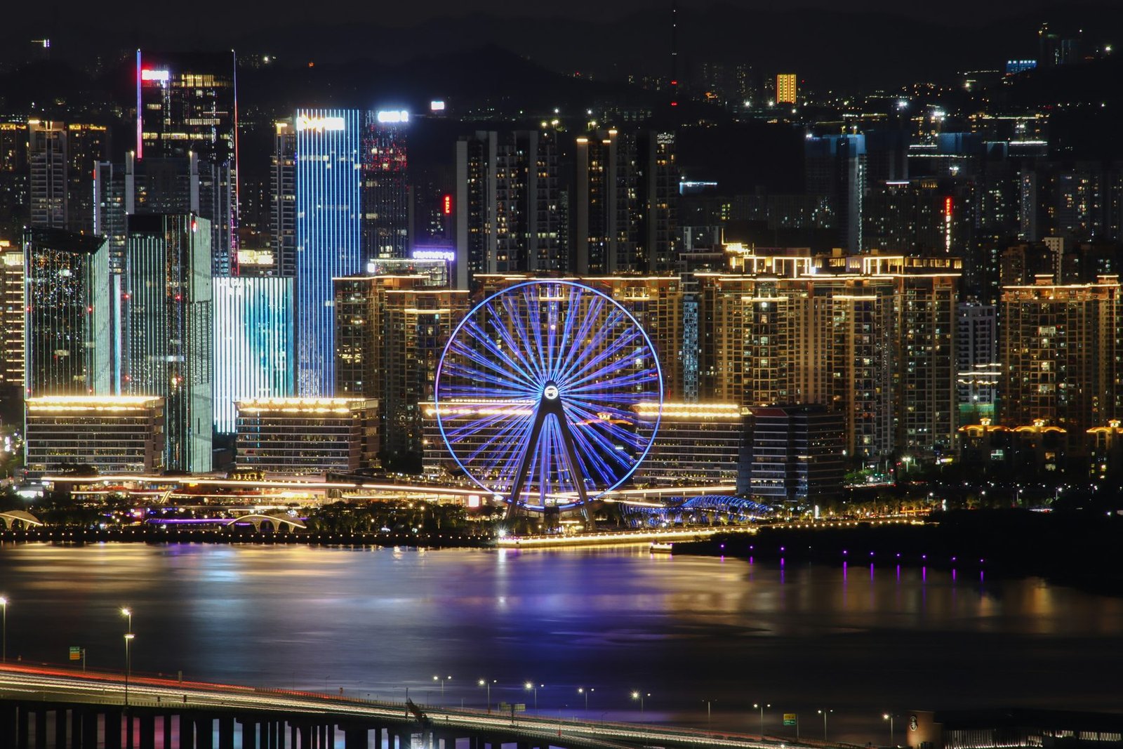 A Ferris wheel lit blue stands among brightly lit towers beside dark water