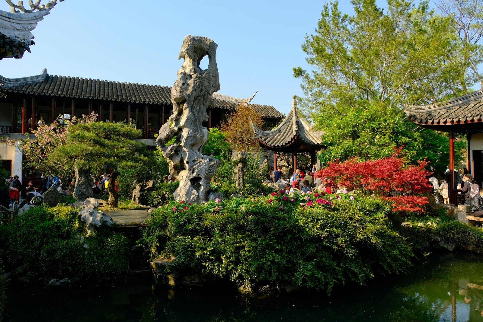 A tall weathered limestone rock stands among trimmed trees and pavilions beside a pond