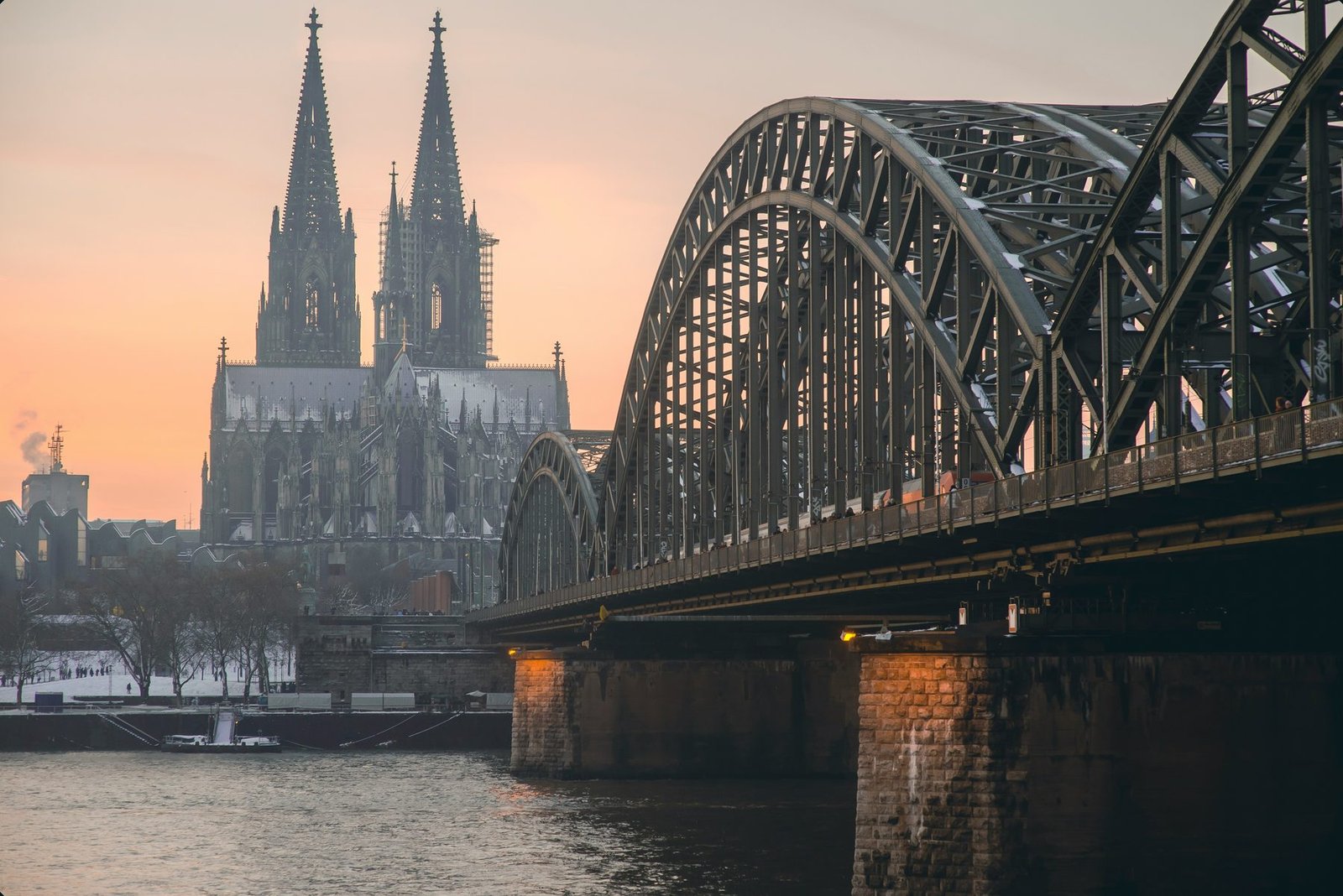 Twin spires stand in soft pink light behind the dark steel arches and stone pier of a railway bridge over a river